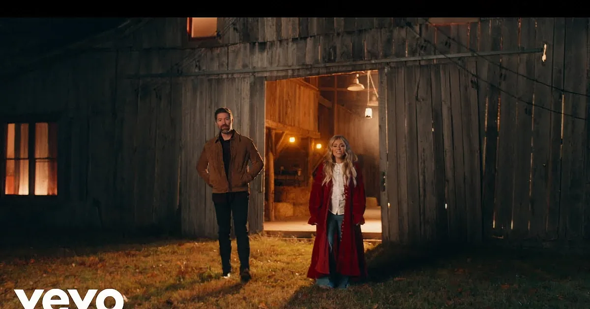 A man and woman stand outside a barn at night, the woman raising her hands as if in surprise or excitement.