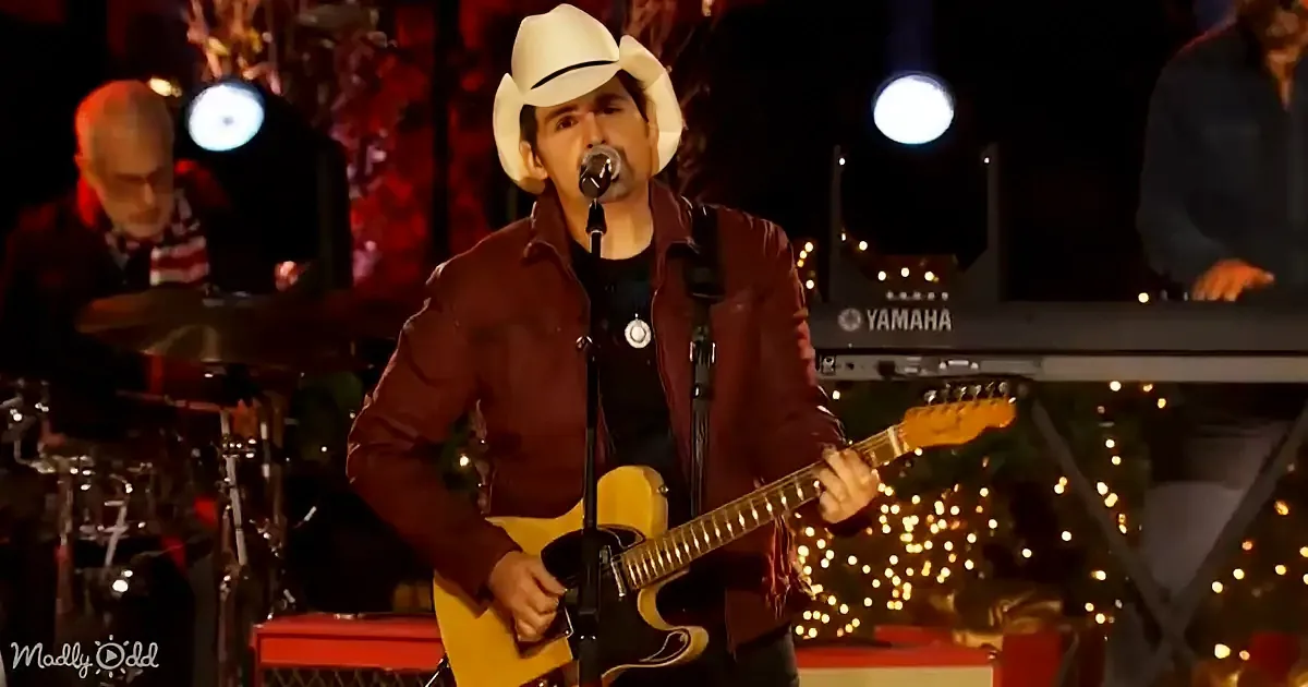 A man in a white cowboy hat plays a yellow guitar on stage with a band and Christmas lights in the background.