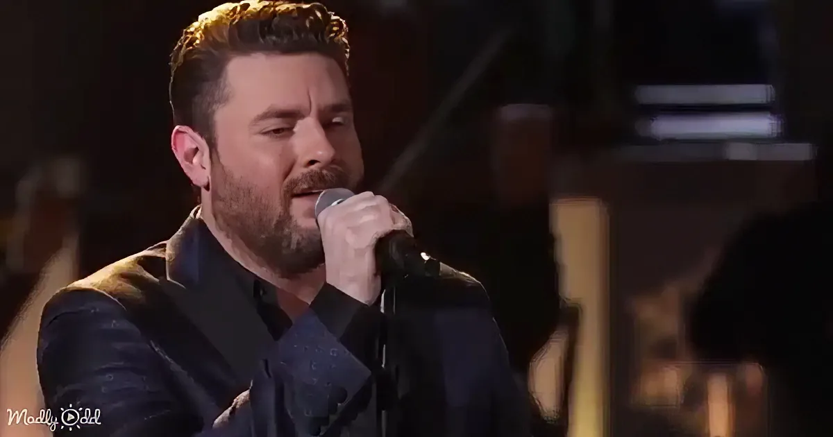 A man with a beard and short hair sings into a microphone, wearing a dark suit jacket.
