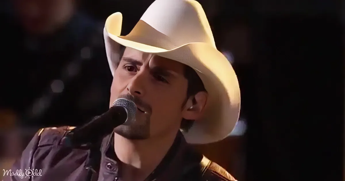 A man wearing a white cowboy hat sings into a microphone, with a dark background and a blurred figure in the top left corner.