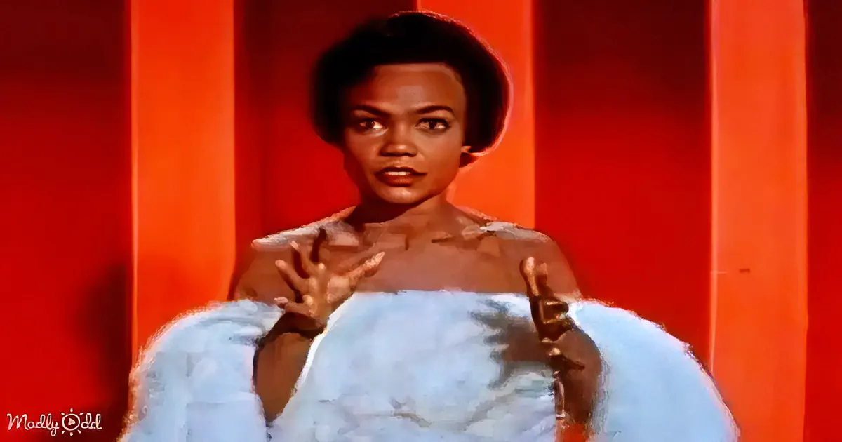 A person in a light blue fur coat gestures with hands, standing against a red-orange striped background.