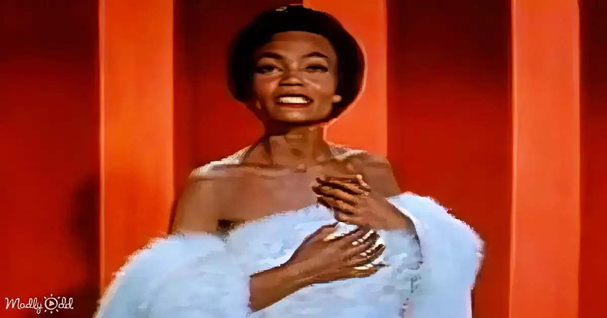 A smiling woman in a light blue dress with a black hat, hands clasped near her chest, against a red-orange background.