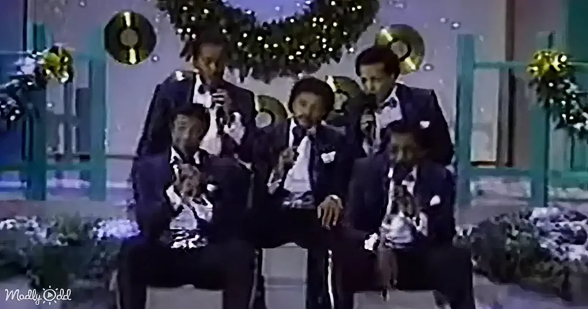 A group of five men in suits sing on stage, holding microphones, with festive decorations in the background.