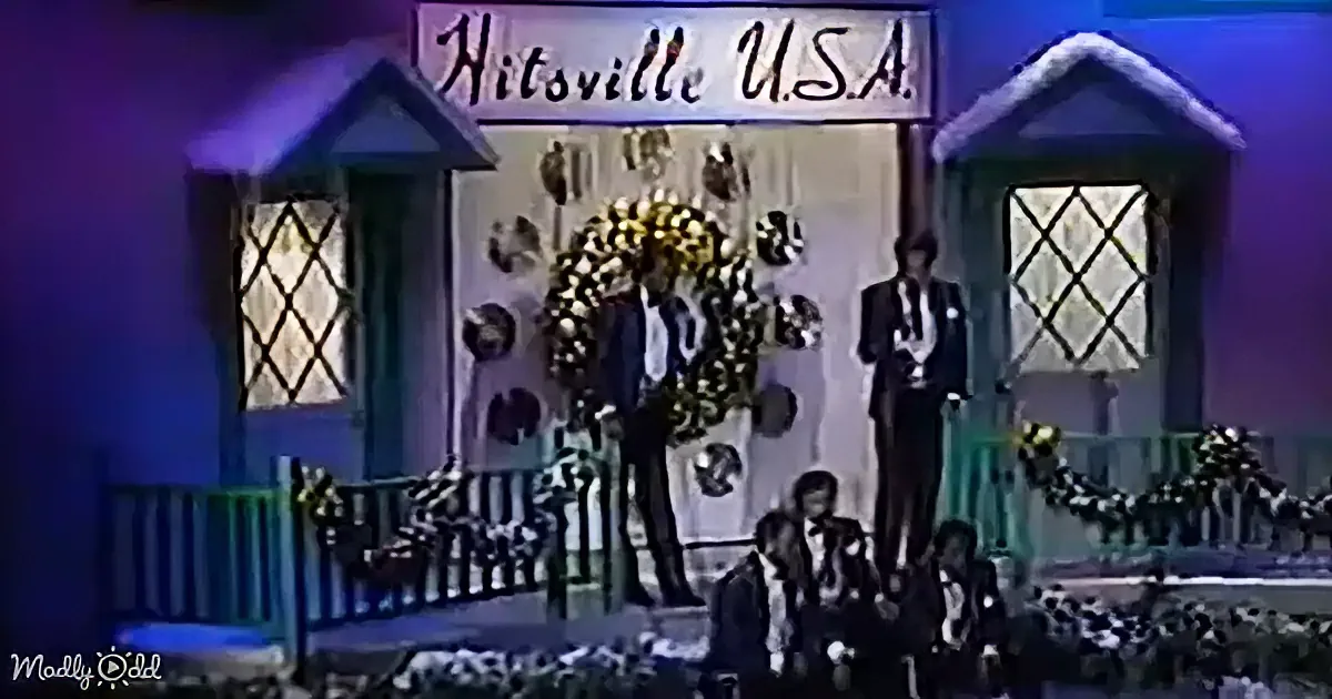 Two men in suits stand on a stage with a "Hitsville U.S.A." sign, surrounded by decorative wreaths and a crowd.