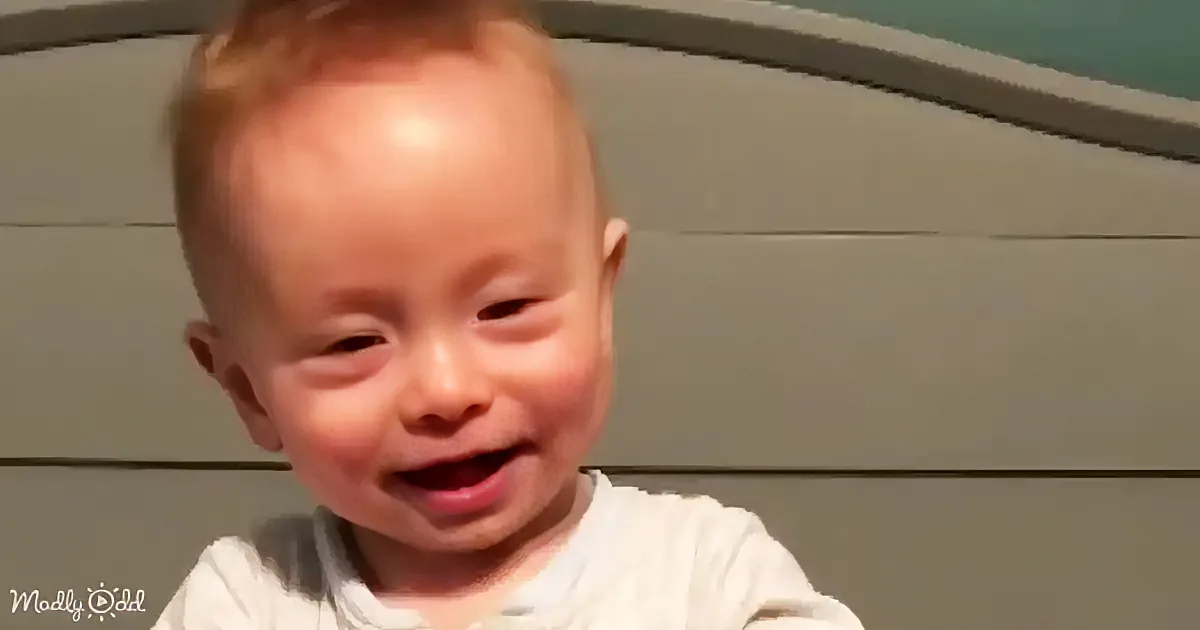 A baby with light skin and short hair is smiling, wearing a white shirt, with a gray background.