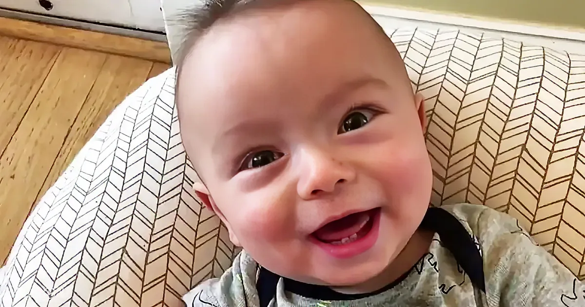 A smiling baby with a light complexion, wearing a patterned shirt, is lying on a white and black patterned surface.