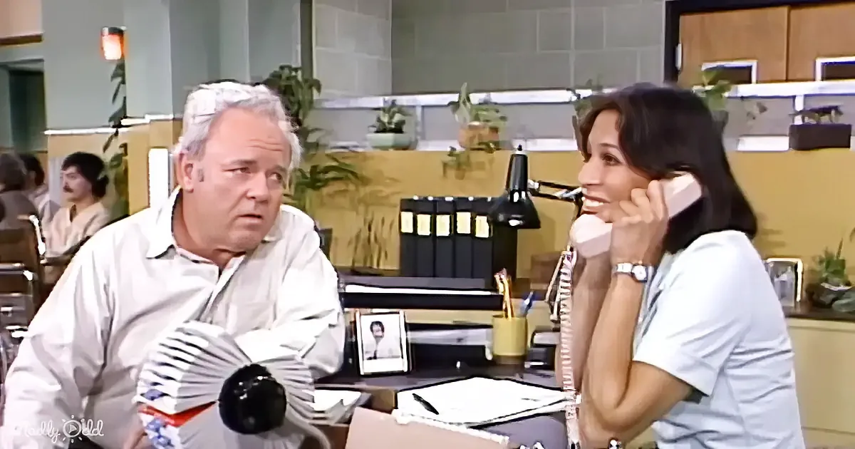 A man and woman are in an office, the man looks surprised while the woman talks on the phone.