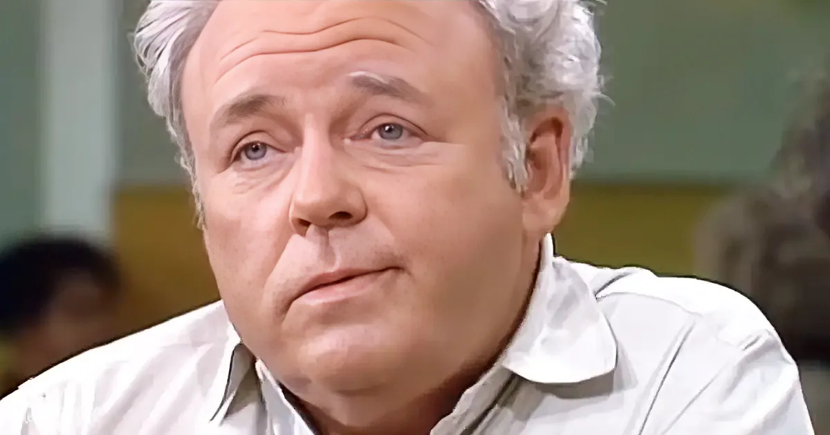 An older man with gray hair, wearing a white shirt, looks to the side with a serious expression.