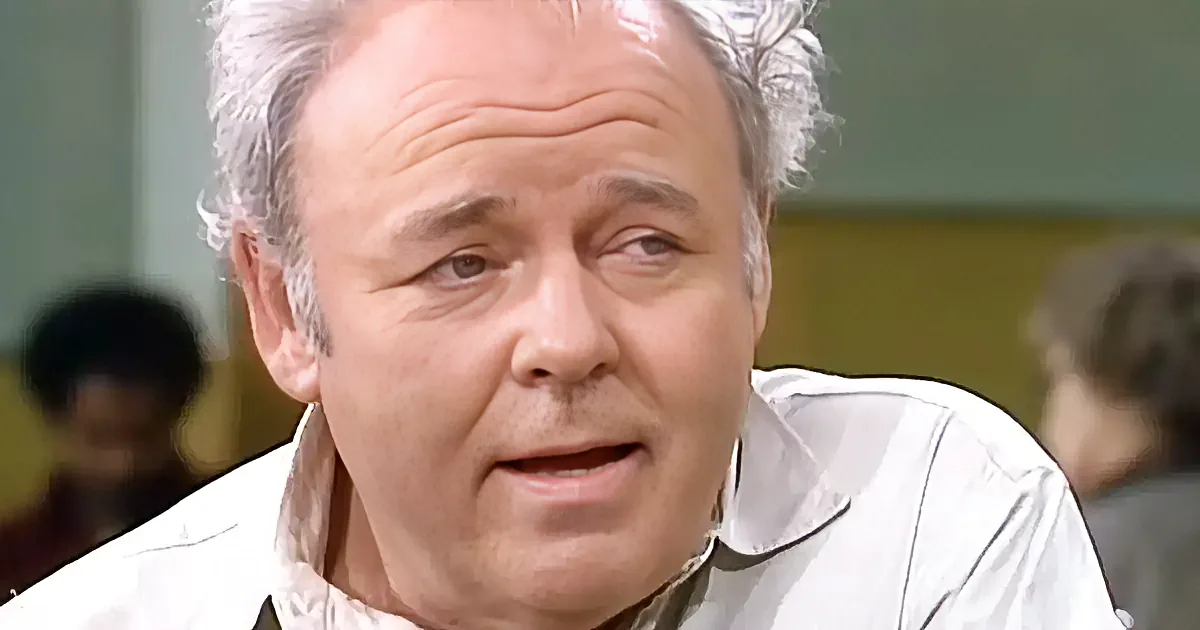 An older man with white hair, wearing a white shirt, is speaking with a slightly open mouth.
