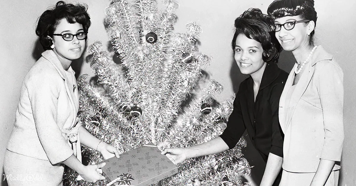 Three women in 1960s attire gather around a tinsel Christmas tree, holding a wrapped gift.