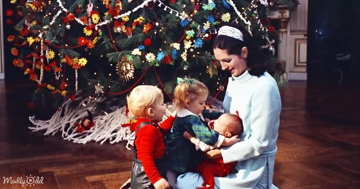 A woman holds a baby while two children look at a decorated Christmas tree.