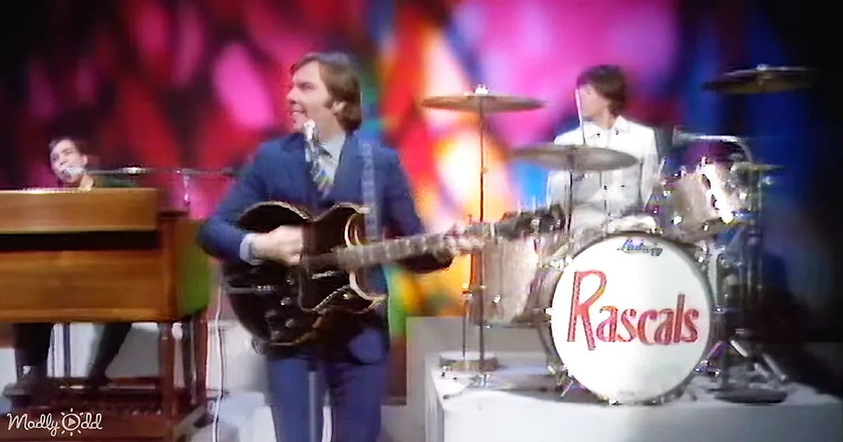A man in a blue suit plays guitar on stage with a colorful background and a drummer behind him.