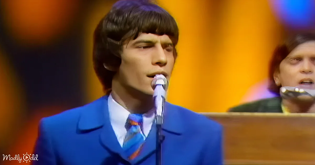 A man in a blue suit sings into a microphone on stage, with a blurred background and another person visible.