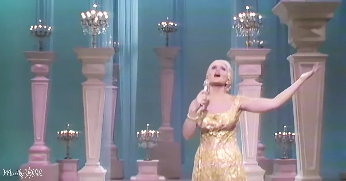 A woman in a gold dress sings on stage, surrounded by chandeliers and columns.