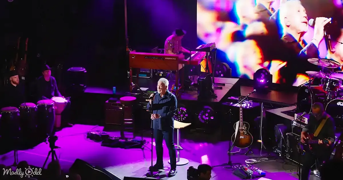 A man in a blue suit sings into a microphone on stage, surrounded by musicians and colorful lighting.