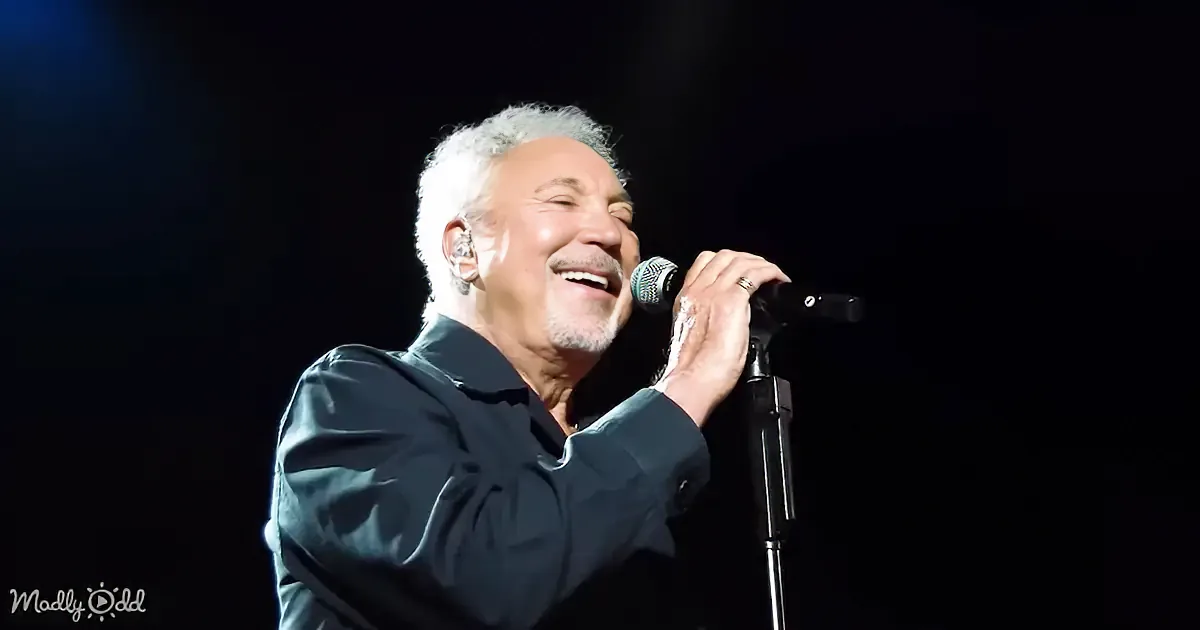 A man with white hair and a beard sings into a microphone on stage.