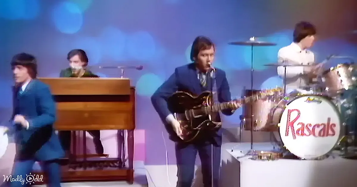 A man in a blue suit plays guitar on stage with a band, including a drummer and keyboardist.