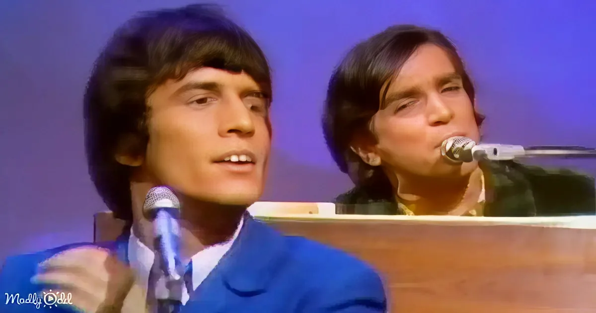 Two individuals in a performance setting, one singing into a microphone, both wearing suits, with a piano in the background.