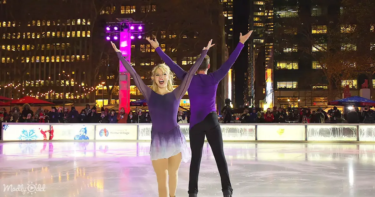 A couple in purple and black ice skating costumes raises their arms in celebration on an ice rink.