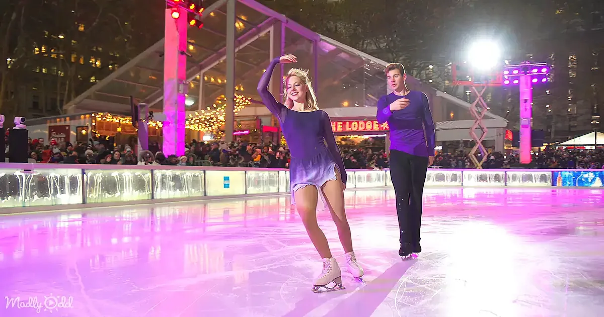 A woman in a purple outfit skates gracefully on an ice rink with a man in a blue shirt, surrounded by festive lights and...