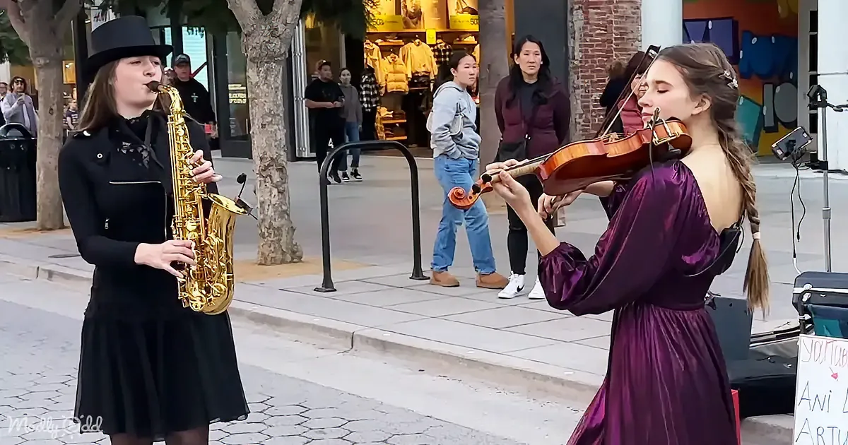 A woman in a black dress plays saxophone while another in a purple dress plays violin on a city sidewalk.