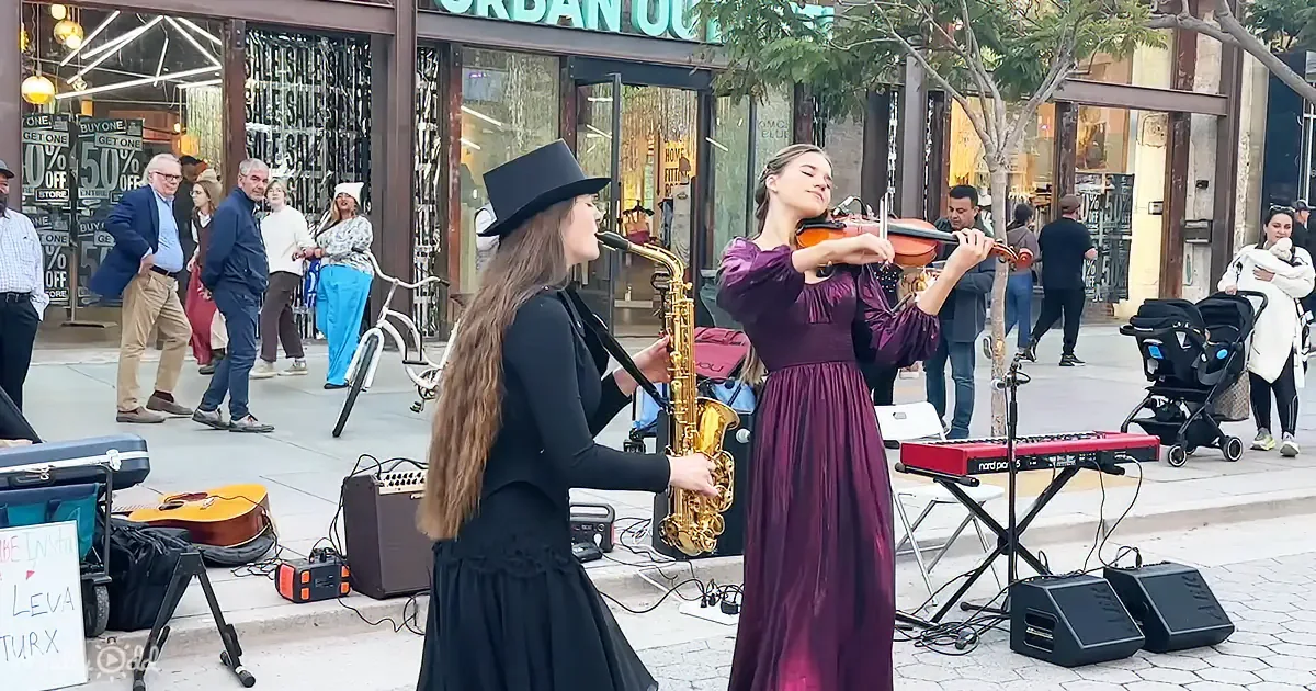A woman in a black dress plays saxophone while another in a purple dress plays violin on a city street.