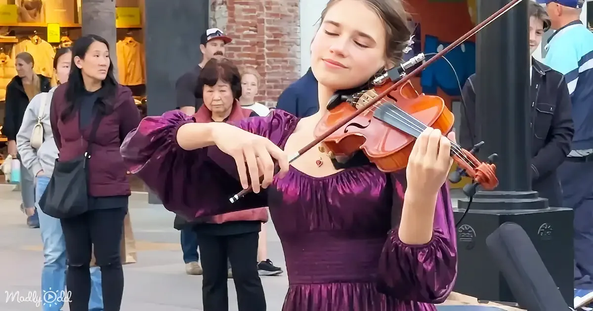 A woman in a purple dress plays the violin on a busy street, surrounded by pedestrians.