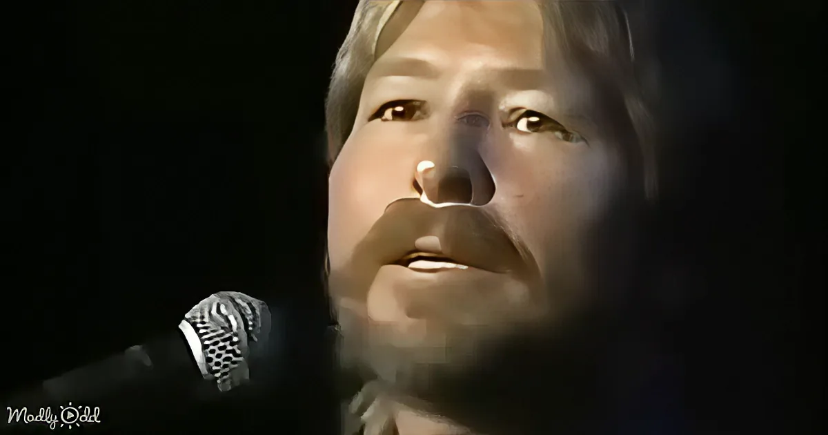 A man with blonde hair and a beard sings into a microphone, his face partially illuminated.