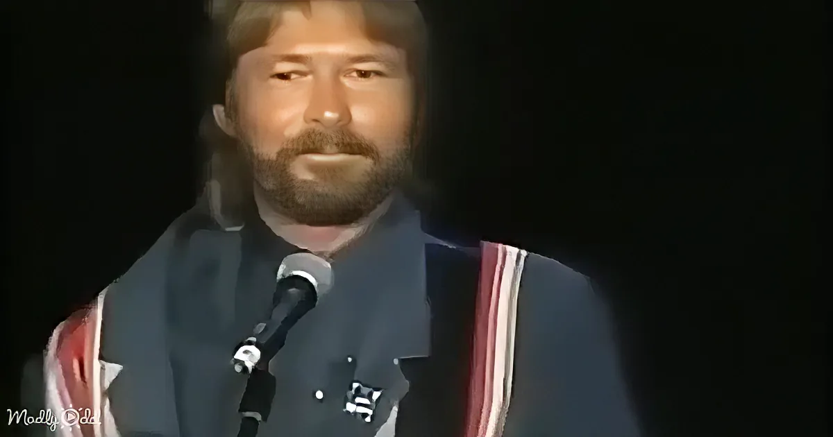 A man with long hair and a beard speaks into a microphone, wearing a dark jacket with red and white stripes.