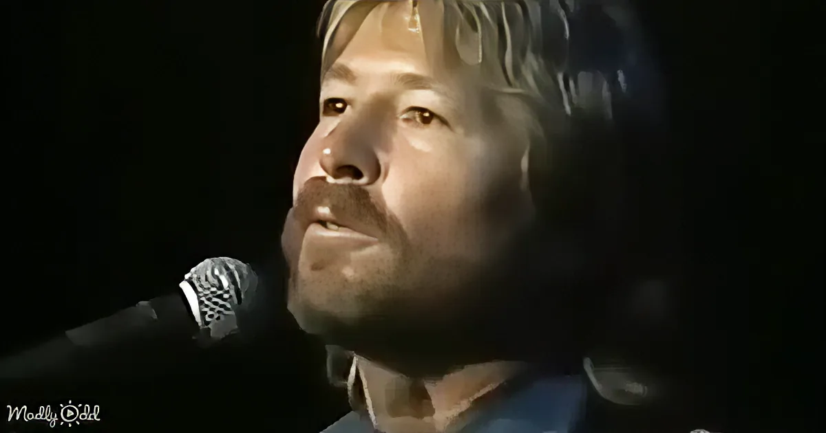 A person singing into a microphone, illuminated by stage lighting, with a dark background.