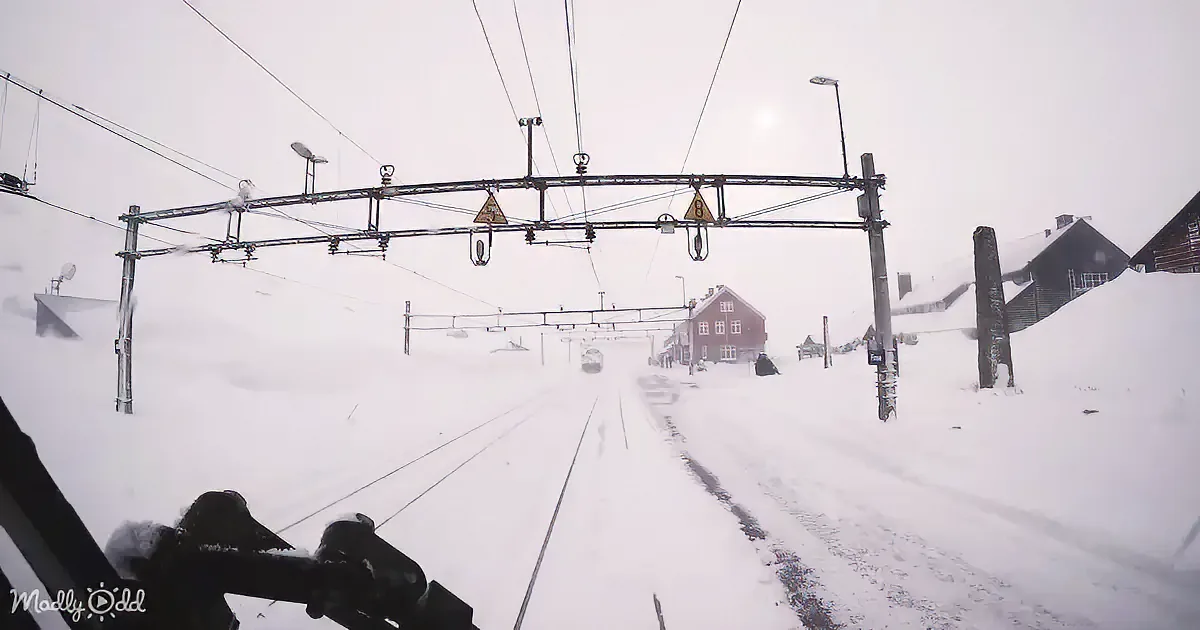 A snowy railway scene with tracks, overhead wires, and a distant train approaching under a cloudy sky.