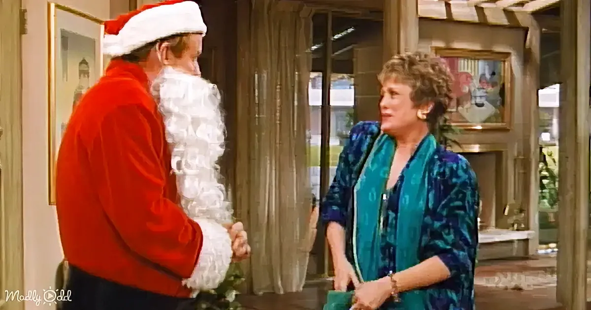 A man in a Santa outfit stands facing a woman in a teal patterned shirt, both in a warmly lit room.