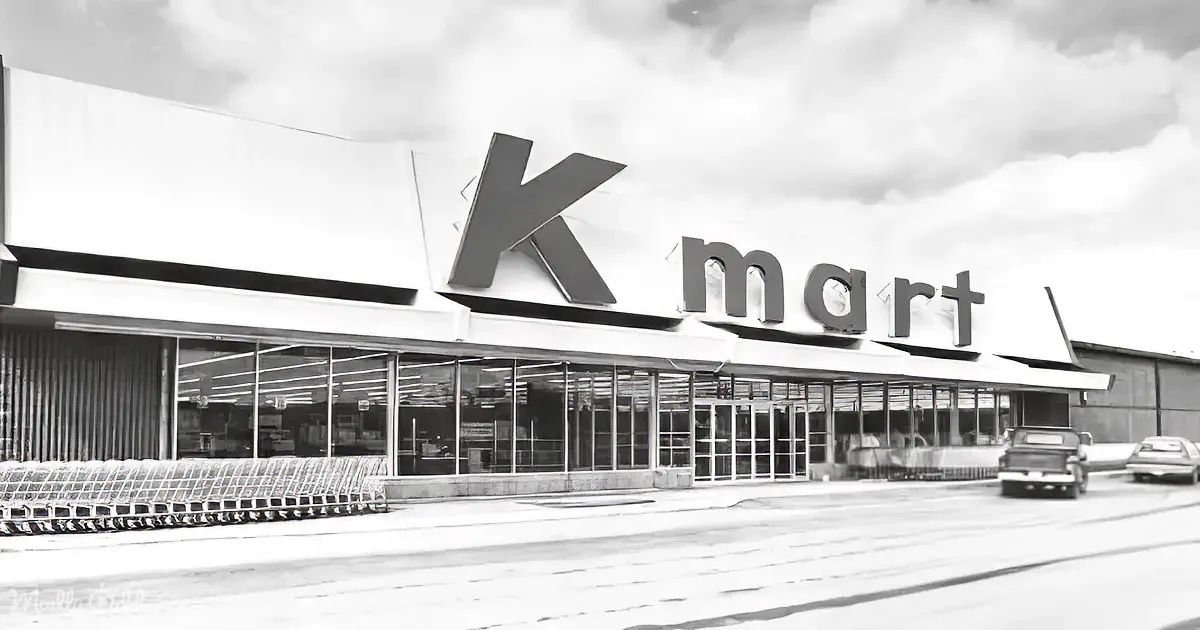 A Kmart store with a large "Kmart" sign, shopping carts, and a cloudy sky in the background.