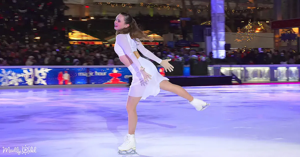 A figure skater in a white dress performs a graceful pose on an ice rink, surrounded by a festive crowd and colorful lights.