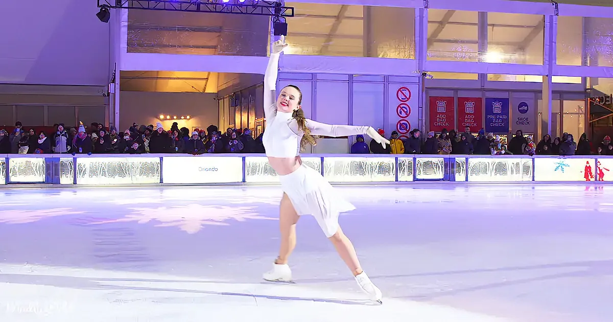 A figure skater in white performs a graceful pose on an ice rink, surrounded by spectators.