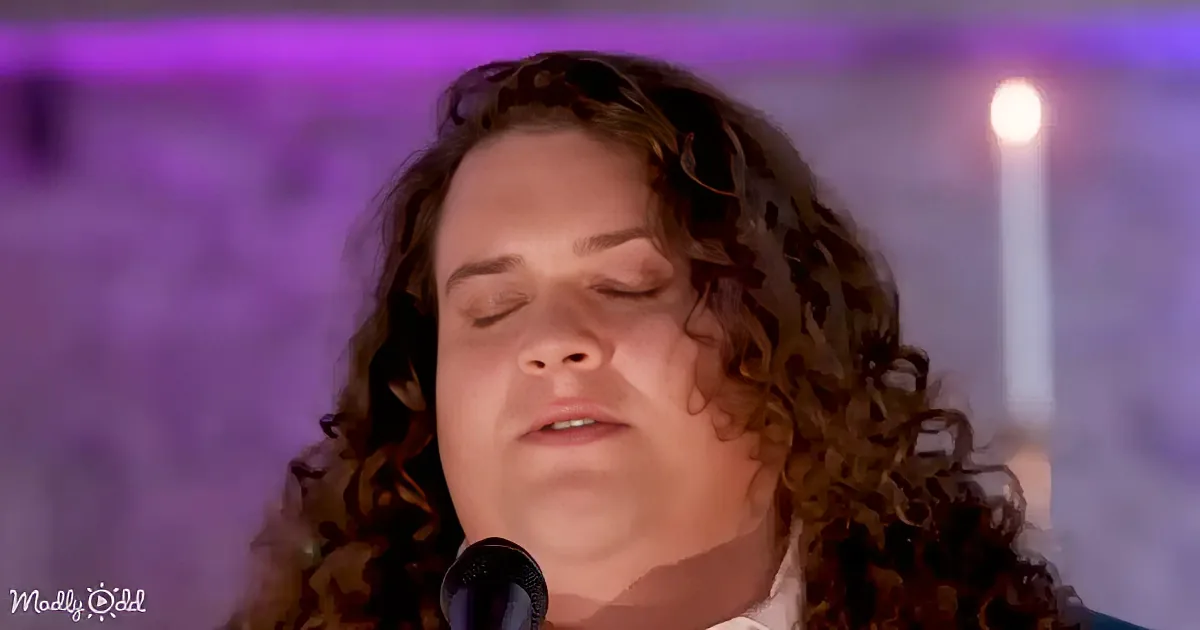 A person with curly hair sings into a microphone, eyes closed, in front of a purple-lit background.