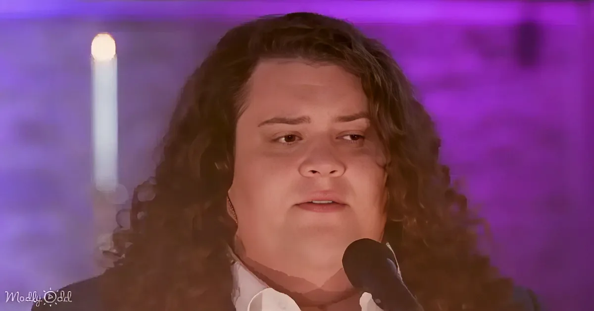 A person with long, curly hair sings into a microphone against a purple-lit background.