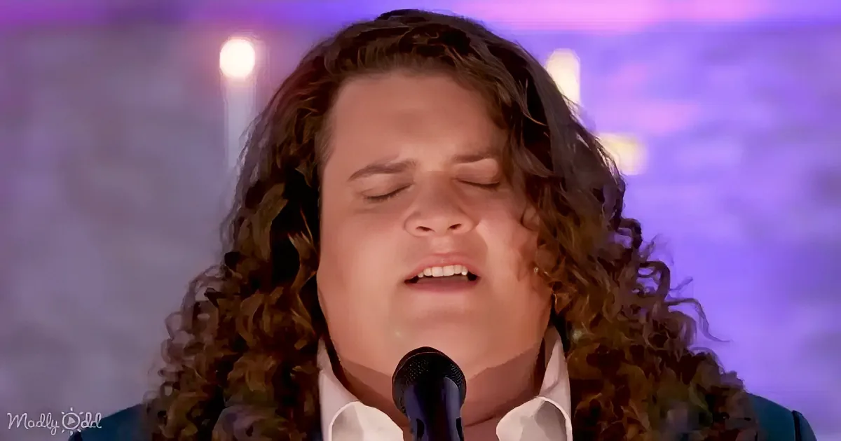 A person with long, curly hair sings into a microphone against a blurred background with purple and white hues.