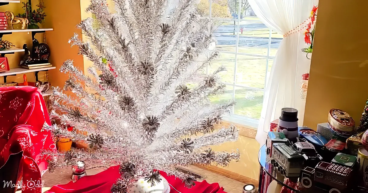 A silver Christmas tree adorned with ornaments sits in a room with a red tablecloth and a window view of a park.