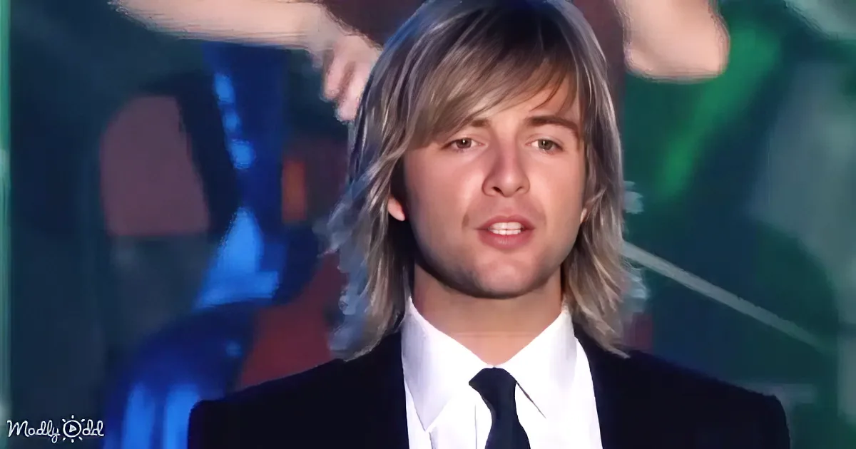 A man with long hair and a suit is speaking on stage, with blurred background and a watermark.