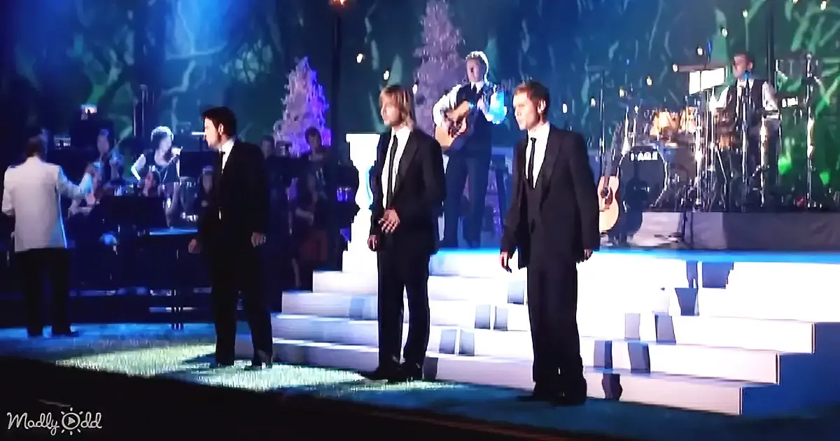 Three men in suits stand on illuminated steps, surrounded by musicians and a festive backdrop.