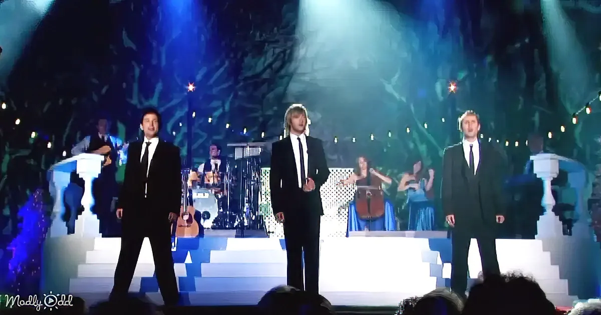 Three men in suits stand on a stage with a band and string quartet in the background.