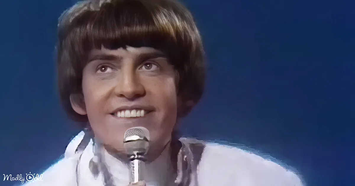 A person with a microphone, wearing a white outfit, against a dark blue background.