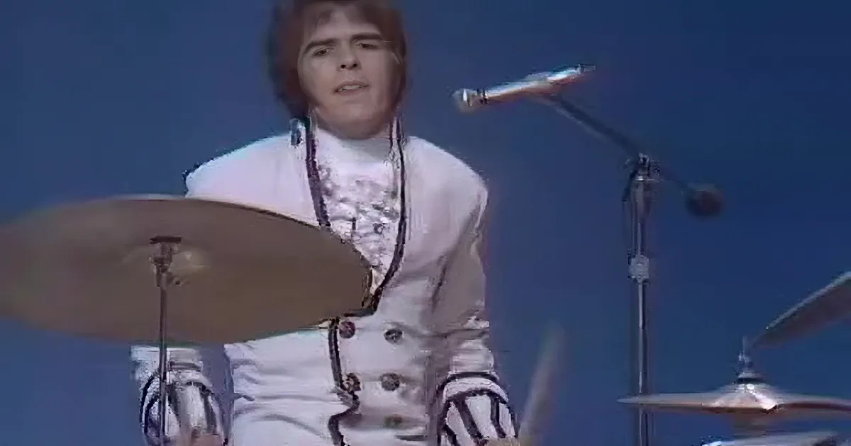 A person in a white outfit sings into a microphone, with another person in the background holding a guitar.