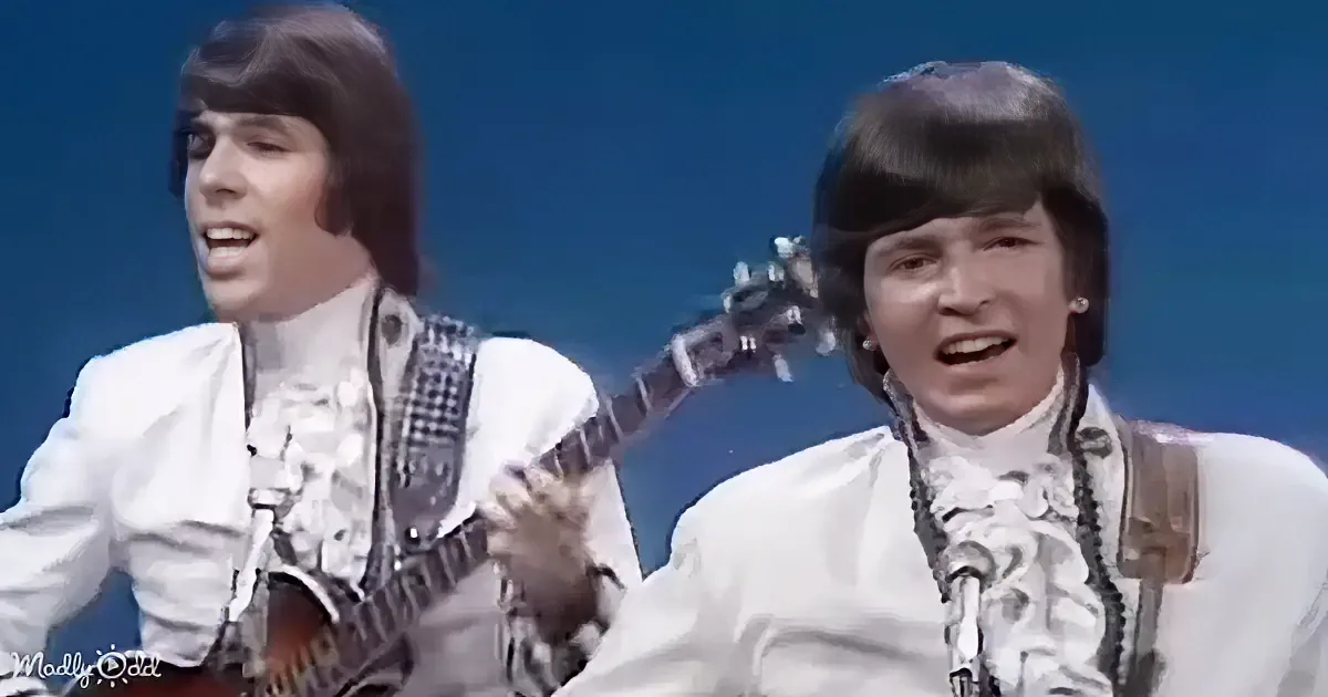 Two individuals in white outfits play guitars and sing on stage against a blue background.