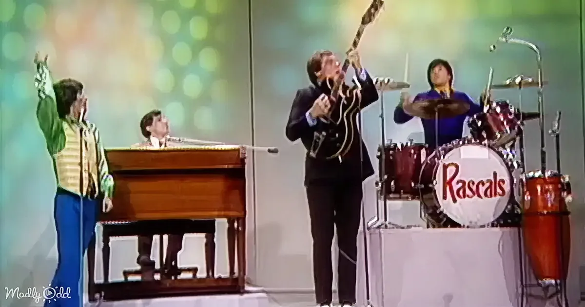 A guitarist in a black suit plays passionately on stage with a colorful background and band members.