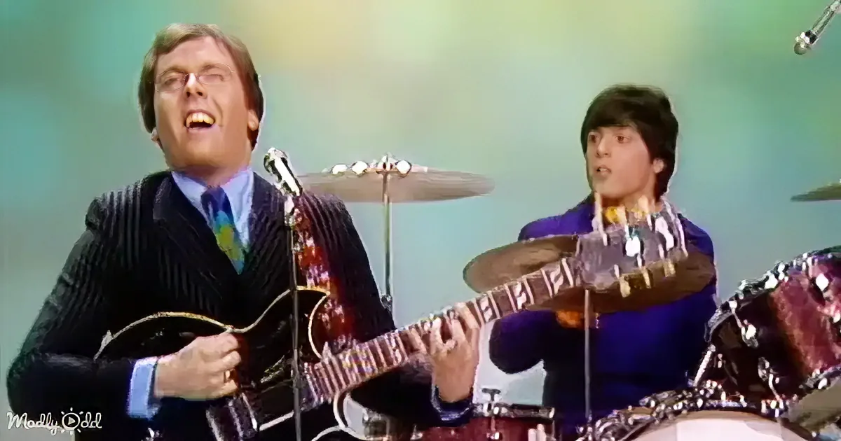 A man in a suit plays guitar while another in a blue shirt plays drums, both singing.