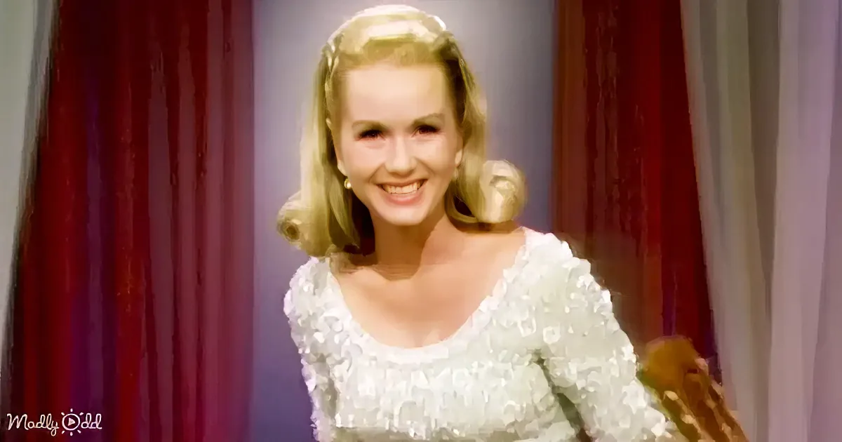 A smiling woman with blonde hair in a white lace dress stands in front of red curtains.