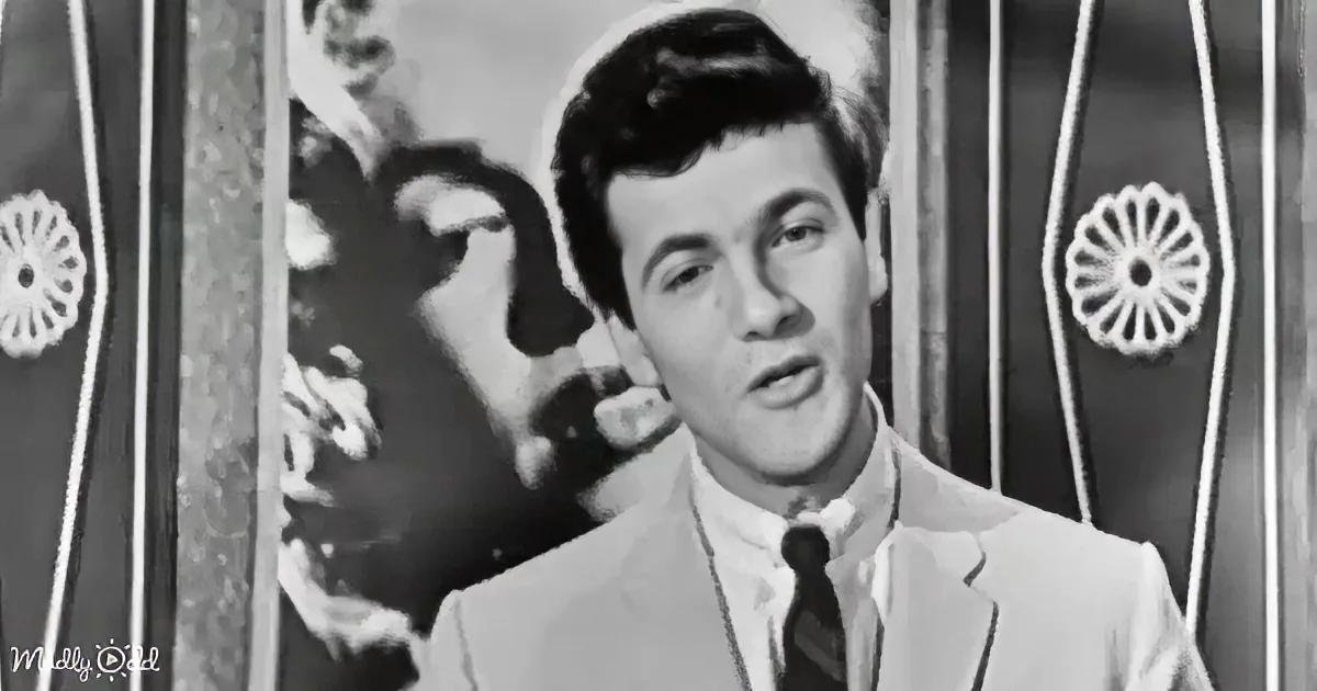 A man in a suit and tie stands in front of a decorative backdrop, appearing to speak or sing.