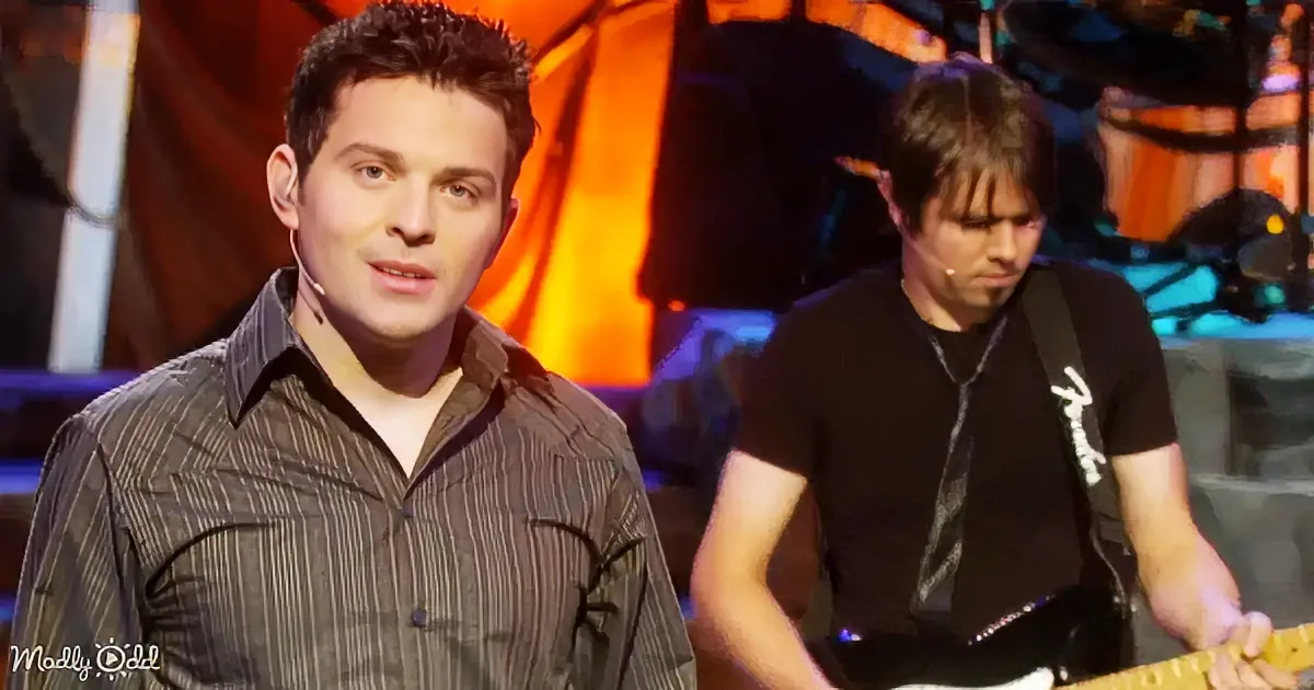 A man in a striped shirt stands on stage, looking to the side, with another man playing guitar behind him.